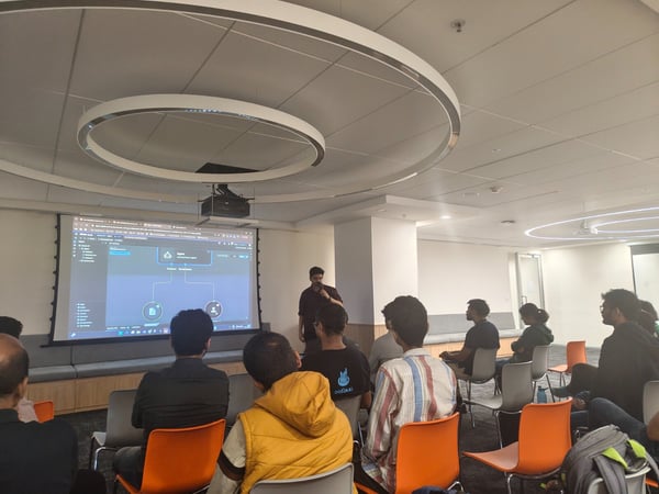 A photograph captures an indoor technical presentation where a speaker stands next to a large projection screen displaying a dark-themed flow chart or application interface, while an audience is seated, watching the demonstration.