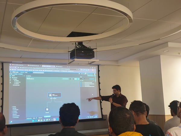 A man is giving a technical presentation, pointing at a software interface (UiPath Studio) projected onto a large screen while speaking into a microphone, observed by an audience seated in the foreground.
