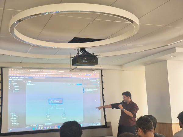 A man is giving a technical presentation or demonstration in a modern meeting room, pointing toward a large projected screen displaying a software interface with diagrams and menus. Several audience members are visible in the foreground, observing the screen.