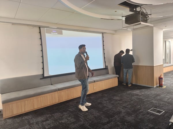 A candid photograph taken inside a modern office or meeting room shows a man holding a microphone and giving a presentation in front of a large projection screen, while two other individuals stand near the wall to the right.