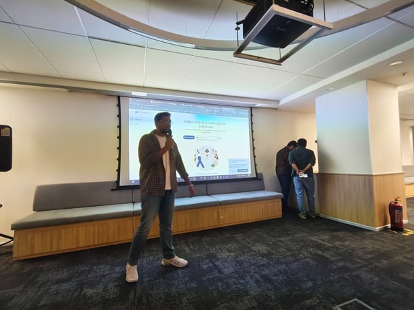 A man stands in an office meeting space holding a microphone and giving a presentation in front of a large projection screen displaying the Google Meet interface and the headline 'Video calls and meetings for everyone.' Two other men are visible standing near a pillar on the right side of the screen.