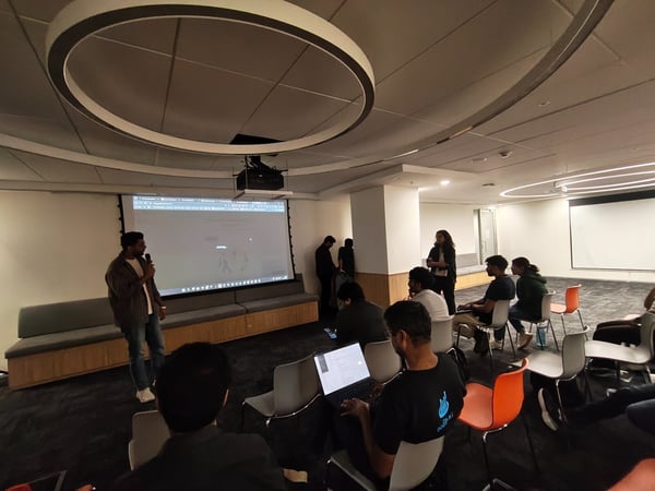 A man is giving a presentation or talk into a microphone in a modern, dimly lit indoor space, while a group of attendees sits and watches the projected screen.