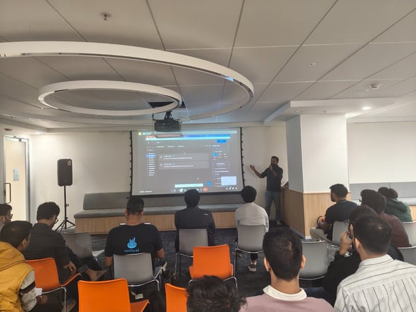 A photograph capturing an indoor presentation where a speaker stands next to a large projection screen displaying a software interface, addressing a seated audience of approximately 18 people.