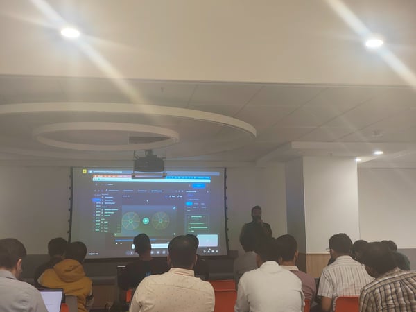 A photo taken indoors shows an audience seated, facing a large projection screen displaying technical content, while a presenter stands to the side giving a talk.