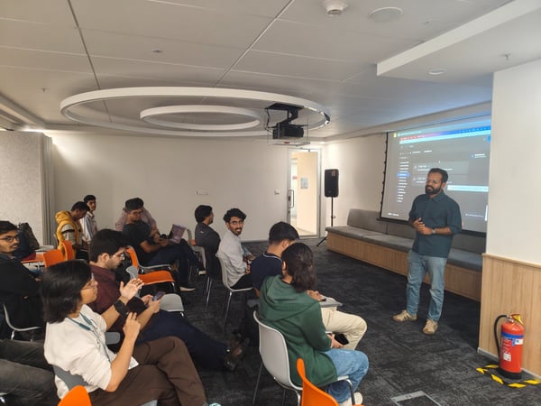 A man gives a technical presentation to a seated audience in a modern office or training room, with content projected onto a large screen behind him.