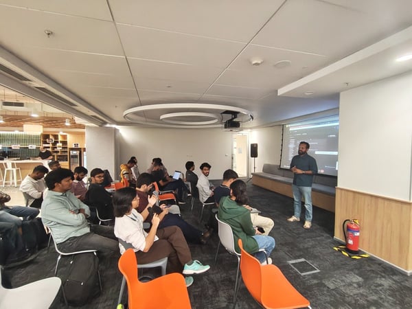A candid photograph capturing an indoor technical presentation or meeting in a modern office space, showing a speaker standing by a large projection screen addressing a seated audience.