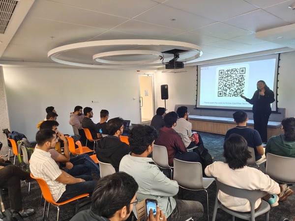A photograph captures an indoor presentation where a woman is speaking into a microphone next to a large projection screen displaying a QR code and the 'CASHFREE PAYMENTS' logo. A large audience, mostly men, is seated in front of her, attending the talk.