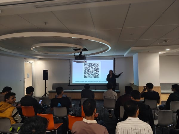 A photograph capturing an indoor tech presentation or seminar, where a speaker stands in front of a projection screen displaying a large QR code and text, addressing an audience seated in rows.