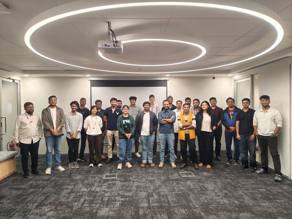 A large group of approximately two dozen young professionals, mostly men, stands together posing for a group photograph in a modern office or conference room.