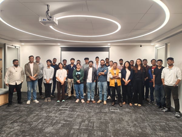 A large group of approximately 26 young professionals, mostly men, stands together for a group photo inside a modern, brightly lit office conference room featuring unique circular ceiling light fixtures.