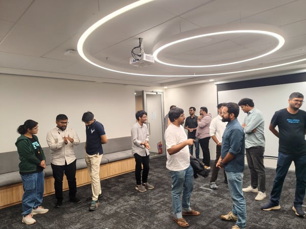 A candid photograph captures a group of approximately a dozen colleagues standing and engaging in informal discussion within a modern office meeting room, characterized by a large circular light fixture on the ceiling.