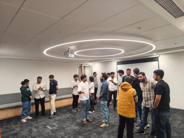 A candid photograph shows a large group of young professionals standing and engaging in conversation in a modern office meeting room, which features a gray carpet, white walls, and distinctive circular ceiling lights.