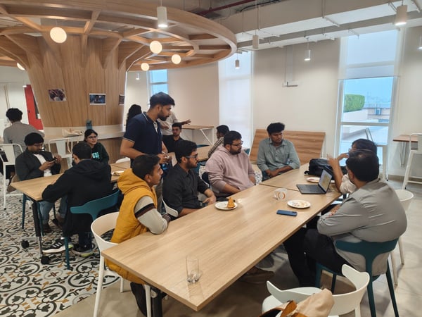 A candid photograph captures a group of young people gathered around wooden tables in a modern, brightly lit office common area or cafeteria, featuring distinctive wooden architectural elements. Several individuals are engaged in discussion, some looking at a laptop, while others are eating snacks.