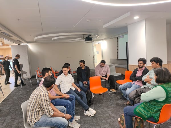 A group of approximately ten people are seated in a circular arrangement on brightly colored orange and gray chairs in a modern indoor setting, likely participating in a discussion or informal team meeting.