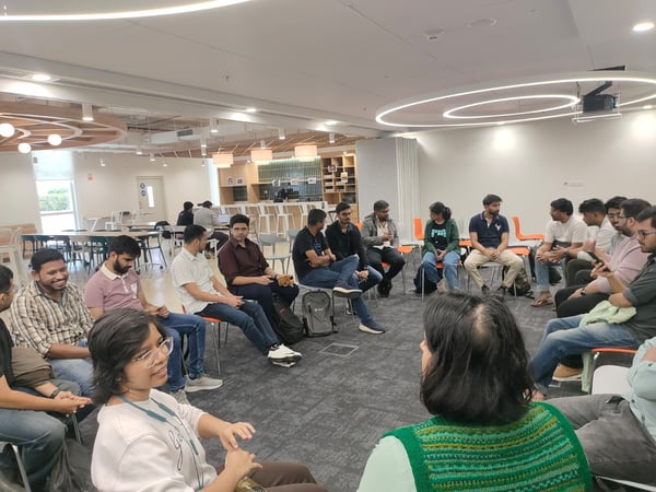A group of approximately seventeen young professionals is seated in a tight circle on chairs inside a modern, brightly lit office lounge or cafeteria area, engaged in an informal discussion.