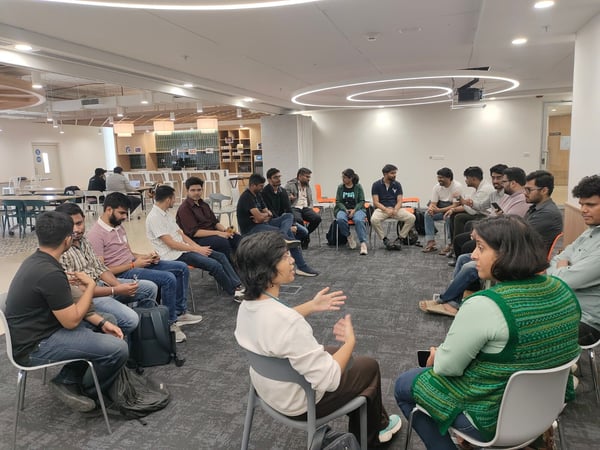 A group of approximately 17 people is seated in a circular arrangement on chairs inside a modern office or co-working space, engaged in a discussion or meeting. Two individuals in the foreground are actively speaking and gesturing towards each other.