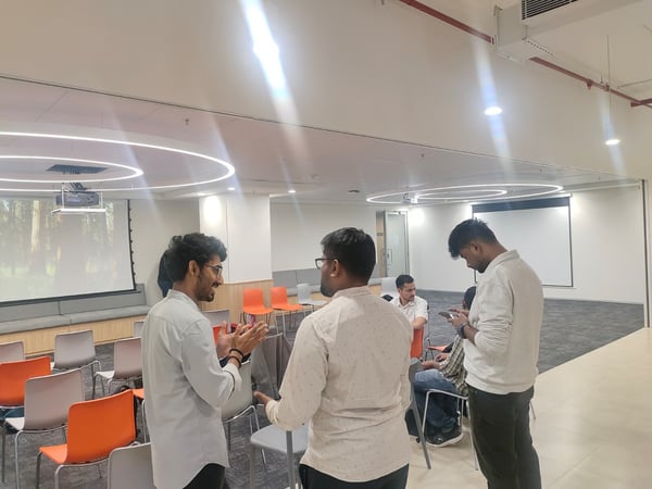 A candid photograph captures a small group of people socializing or networking in a modern, brightly lit indoor conference or event space, featuring rows of empty orange and grey chairs and a projection screen displaying a nature scene.
