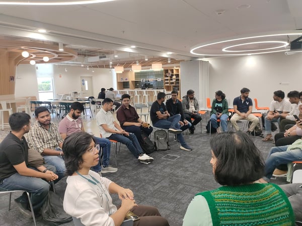 A group of approximately fourteen people is sitting in a tight circle on chairs in a modern, well-lit office lounge or common area, engaged in a group discussion or meeting.