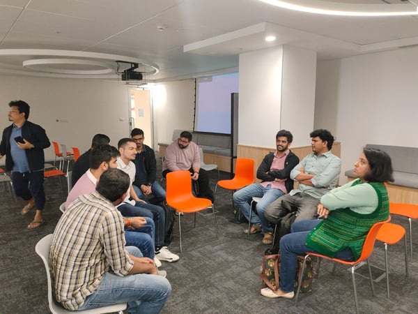 A candid photograph capturing a group of approximately ten people sitting in a semi-circle on orange and white chairs in a modern, brightly lit office meeting room, engaged in a discussion.