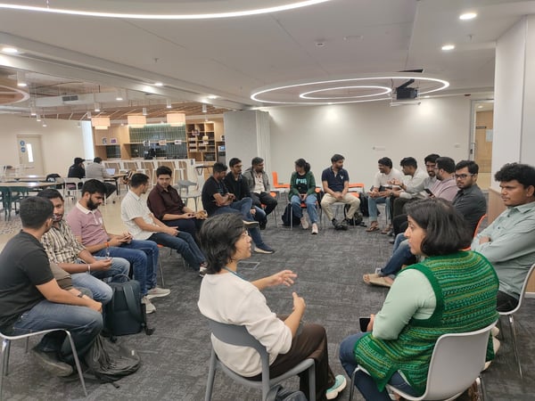 A group of approximately 16 people is seated in a circle on chairs in a modern, open-plan office collaboration area, engaged in a focused discussion or meeting. Two women in the foreground are actively speaking and gesturing toward the group.