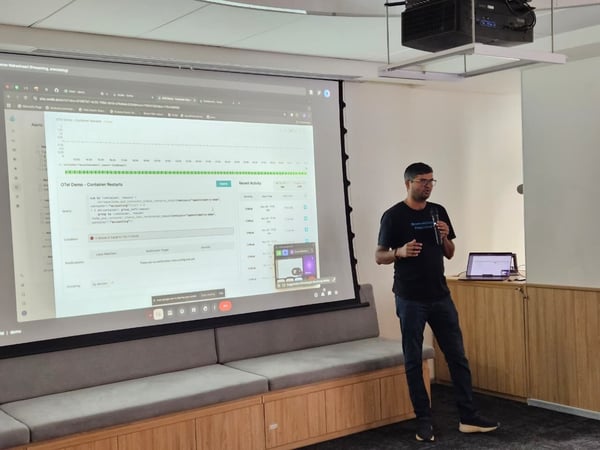 A man wearing glasses and a dark t-shirt is giving a technical presentation or demo in an indoor setting, standing beside a large projection screen that displays a dashboard related to container restarts.