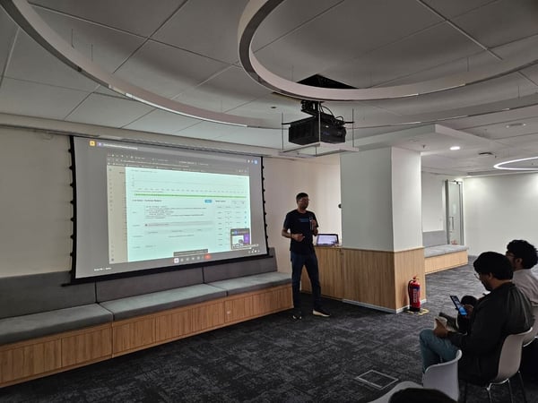 A photograph captures a business presentation taking place in a modern office or conference room, where a presenter stands next to a large projection screen displaying data, while several audience members watch and use their phones or laptops.