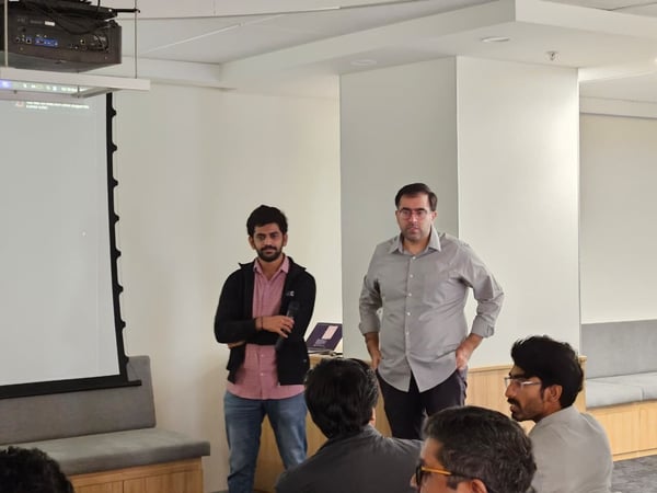 Two men are standing in a modern, well-lit conference room in front of a large projection screen, addressing a seated audience whose heads are visible in the foreground.