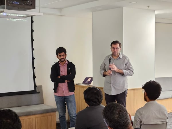 A candid photograph captures two men presenting in a bright indoor setting, with one speaker holding a microphone while several audience members are visible in the foreground with their backs to the camera.