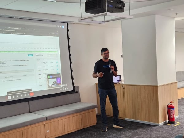 A man wearing a black t-shirt and glasses is giving a technical presentation in a modern office or meeting room, standing next to a large projection screen displaying software monitoring data.