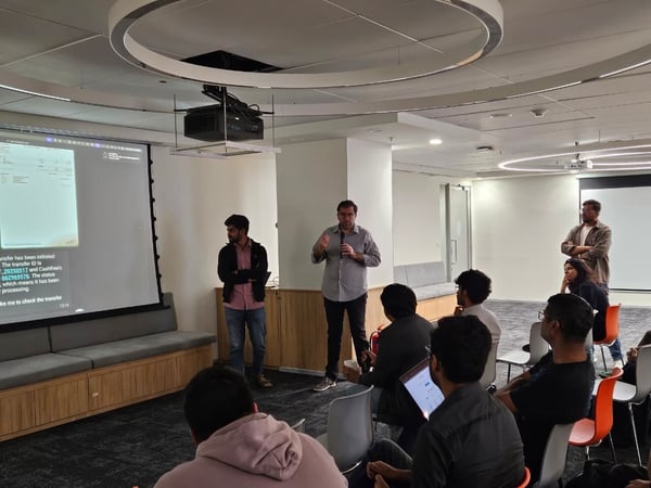 A photograph captures a technical presentation or demo being held in a modern, brightly lit office space, featuring a speaker holding a microphone near a projection screen displaying transaction status details to an audience seated in the foreground.