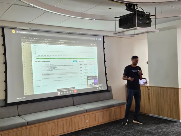 A man wearing glasses and a dark t-shirt stands in a modern meeting room, giving a technical presentation displayed on a large projection screen showing a software dashboard related to container monitoring.
