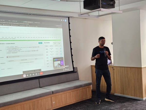 A man wearing glasses and a black t-shirt is giving a technical presentation indoors, standing next to a large projection screen displaying a software dashboard related to container restarts and recent activity.