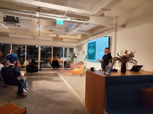 A candid photograph captures an indoor presentation where a speaker stands next to a large illuminated screen displaying content related to a "Worldwide Hackathon," addressing a small, casually seated audience.