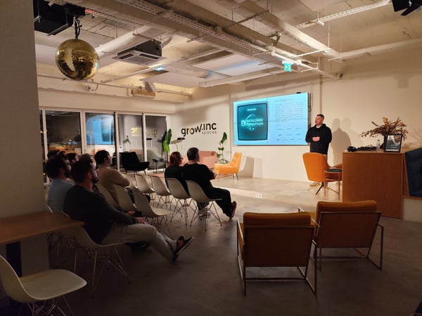 A photograph captures an evening presentation or seminar in a modern, industrial-style event space, where a presenter stands next to a large screen displaying a slide about a 'WORLDWIDE INNOGATHON' to a seated audience.