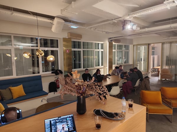 This is a photograph of a modern, stylish indoor co-working or meeting space where several individuals are seated around a long table, primarily engaged in work on laptops. The room is decorated with contemporary furniture, including yellow velvet chairs, a blue built-in sofa, and a striking brass light fixture.
