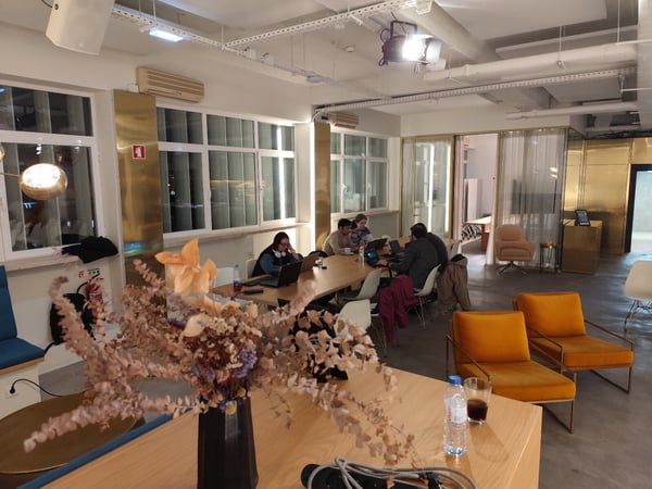 A candid photograph captures five people working on laptops around a long wooden table in a modern, stylish co-working space featuring striking brass wall accents and large windows, with dried flowers visible in the foreground.