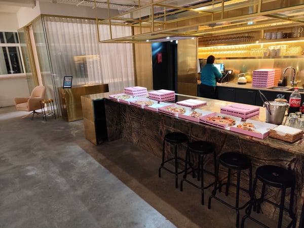 A photograph capturing a modern, stylish indoor bar or catering area featuring a dark marble counter, gold/brass fixtures, and several pink pizza boxes containing pizza slices. One person wearing a teal shirt is visible working behind the counter in the background.