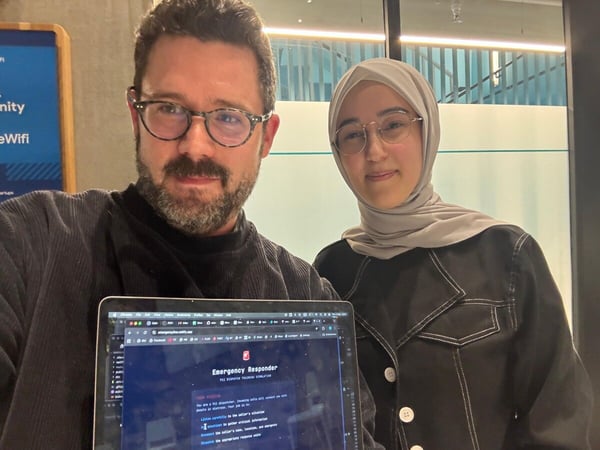 A man and a woman, both wearing glasses, pose for a photo while holding up a laptop that displays a dark-themed application titled "Emergency Responder: 911 Dispatch Training Simulation."