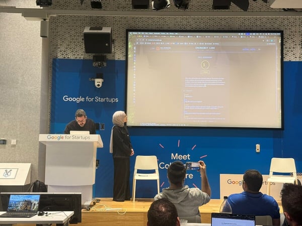 A photograph capturing an indoor technology presentation or pitch event, showing a man at a podium and a woman standing nearby on a stage, facing a large screen displaying a dark-mode web application interface labeled 'EMERGENCY LINE'. Several attendees are visible in the foreground, with one holding a phone to take a picture.