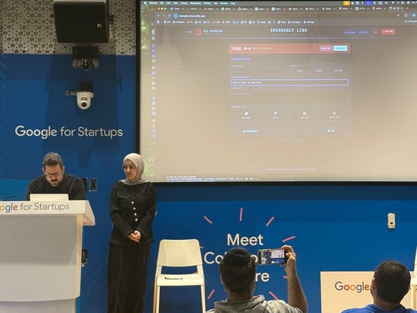 A photograph capturing an indoor tech event or presentation, showing two presenters near a podium and a large screen displaying a web application interface for an 'Emergency Line' dispatch system, with audience members visible in the foreground.