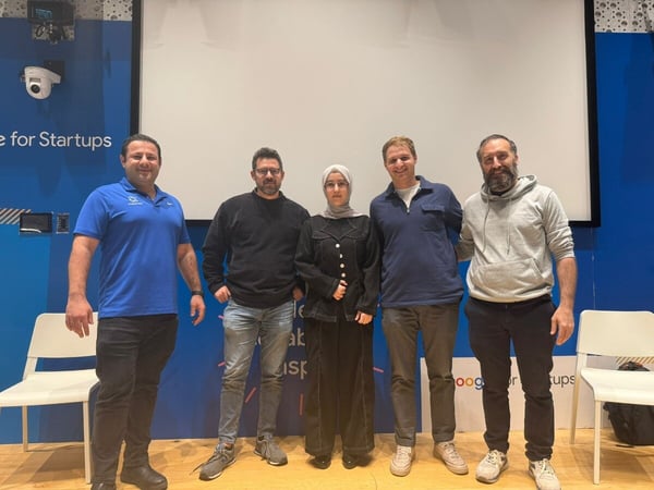 Five individuals, including four men and one woman wearing a hijab, stand side-by-side posing for a group photo in an indoor event space featuring a large white screen and a blue wall branded with 'for Startups' text.