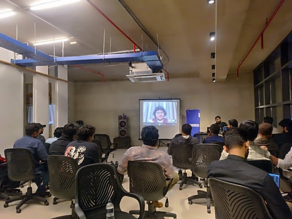 A photograph captures a group of people attending an indoor presentation or seminar, seated in office chairs facing a screen that displays a person via video conference, with a physical presenter standing nearby.