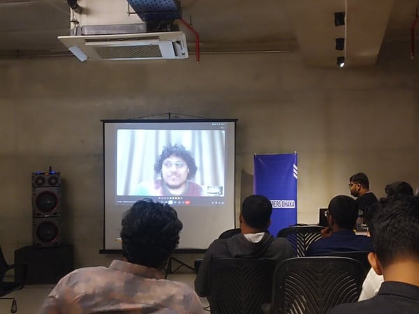 A group of attendees is seated in an indoor setting, watching a virtual speaker projected onto a large screen during what appears to be a presentation or meeting.