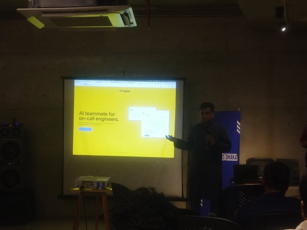 A dimly lit photograph captures a technical presentation where a speaker, dressed in dark clothing, stands next to a large projection screen displaying text about an 'AI teammate for on-call engineers' on a bright yellow background.