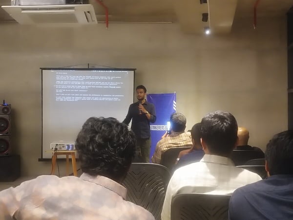 A photograph captures an indoor presentation where a male speaker stands next to a large projection screen displaying text, addressing an audience seated in the foreground, viewed primarily from behind.