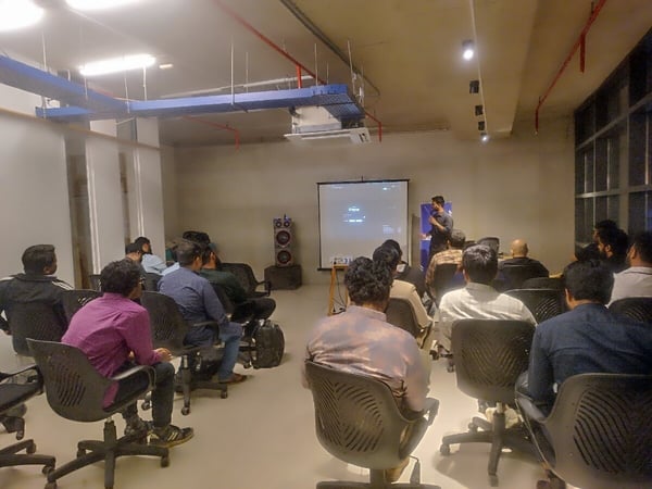 A photograph captures an indoor presentation or seminar where a speaker addresses an audience seated in office chairs, with content displayed on a large, dimly lit projection screen.