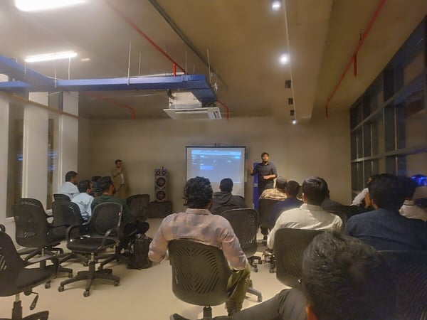 A low-light indoor photograph captures a technical presentation or workshop where a speaker addresses an audience seated in black office chairs facing a projection screen displaying interface elements.