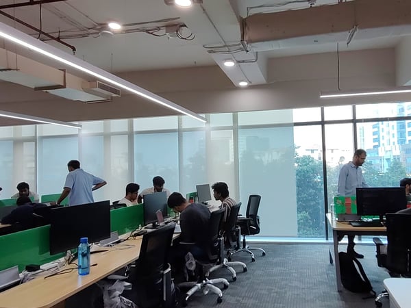 A wide-angle photograph of a modern indoor office space with multiple people working at desks equipped with computer monitors and green privacy dividers.