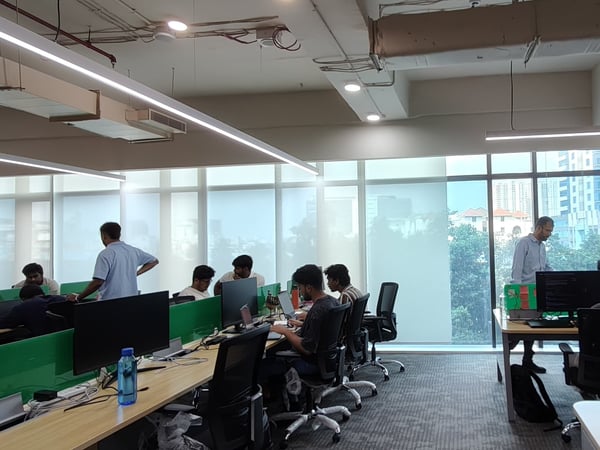 A wide-angle photograph of a modern open-plan office with several employees working at desks equipped with computers and green privacy screens.