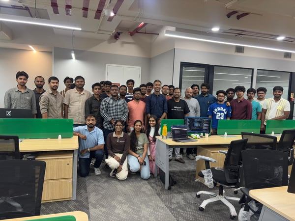 A group photo of approximately 30 people, mostly men, posing together in a modern office setting with desks and computer equipment.